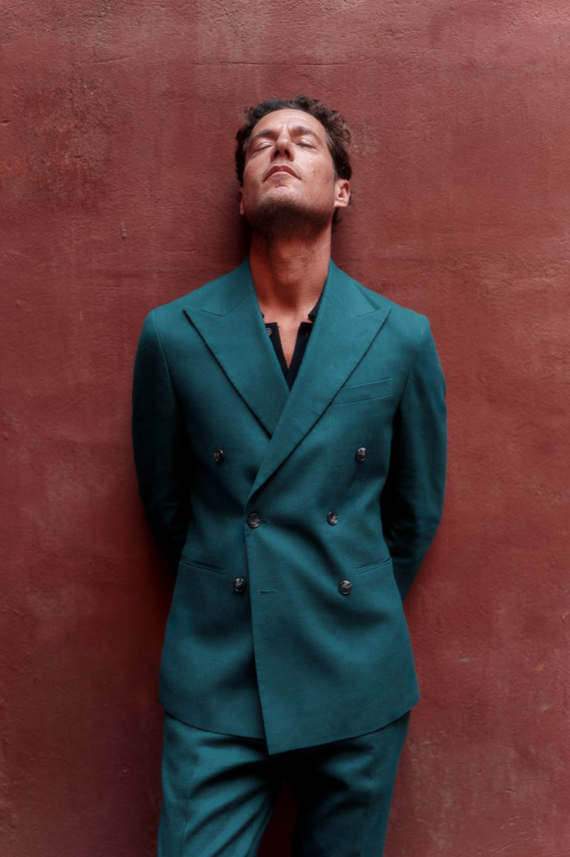 An adult male in a teal suit stands against a red-brown wall with his eyes closed. The image evokes a sense of relaxed sophistication.
