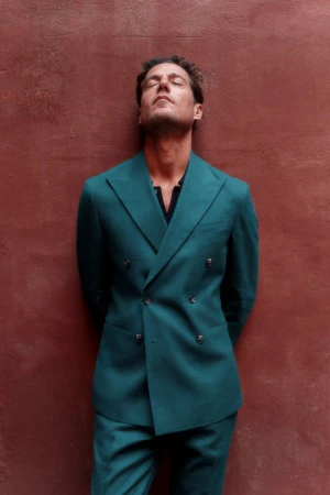 An adult male in a teal suit stands against a red-brown wall with his eyes closed. The image evokes a sense of relaxed sophistication.