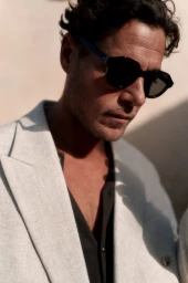 Man wearing dark sunglasses and a light-colored blazer looking down in a sunlit outdoor portrait.