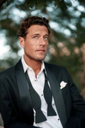 Man in black tuxedo jacket and white shirt with untied bow tie posing outdoors beneath foliage