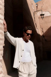Man wearing sunglasses and a white cardigan leaning against a textured stucco wall in an alleyway