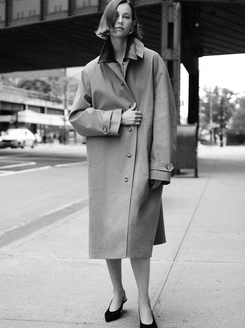 Monochrome shot of a stylish woman in an oversized coat on a city street, standing confidently against an urban backdrop.