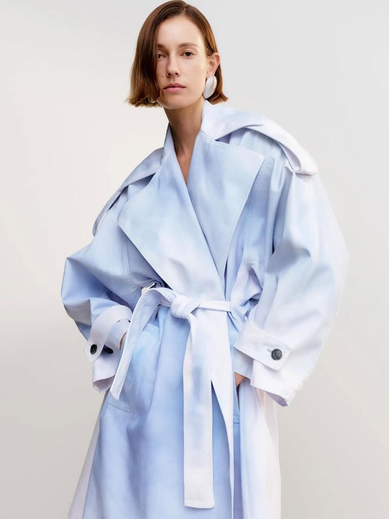 A woman with short brown hair models a blue-and-white ombre belted trench coat in a clean, neutral indoor setting. Fashion editorial shot.