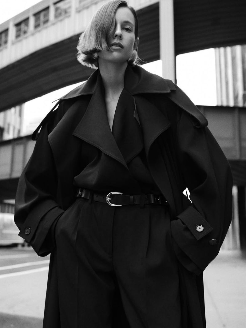 Monochrome fashion portrait of a woman in a dark coat and trousers, standing on an urban street with a bridge in the background.