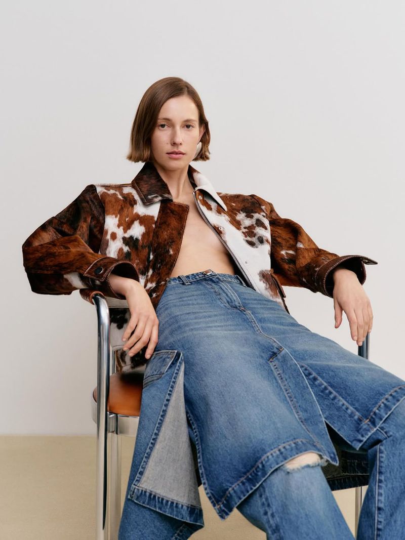 Adult female model in a cow-print jacket and blue denim skirt-pants sits in a metal chair against a white backdrop in a studio.