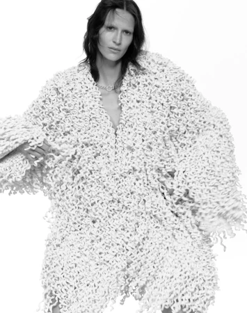 Monochrome fashion shot of a slim adult woman in an oversized fuzzy white coat, gazing directly at the camera against a white background.