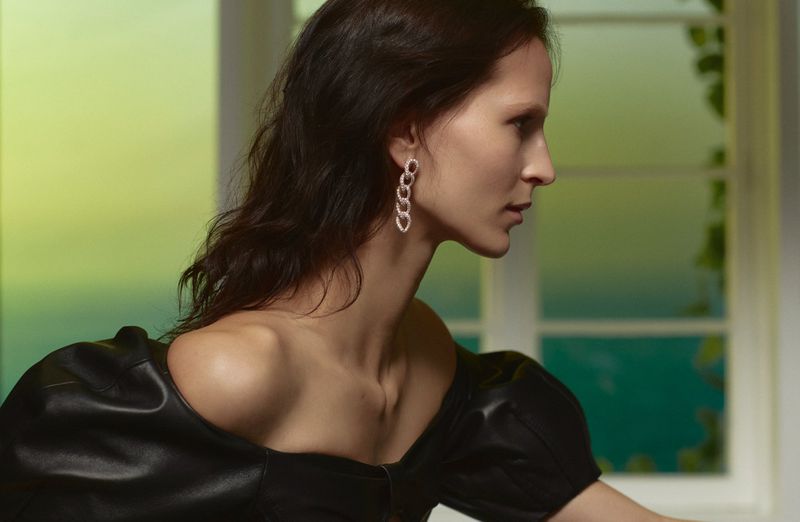 Elegant brunette woman poses in close-up, wearing earrings, with a window and greenery in the background, and soft, diffused lighting.