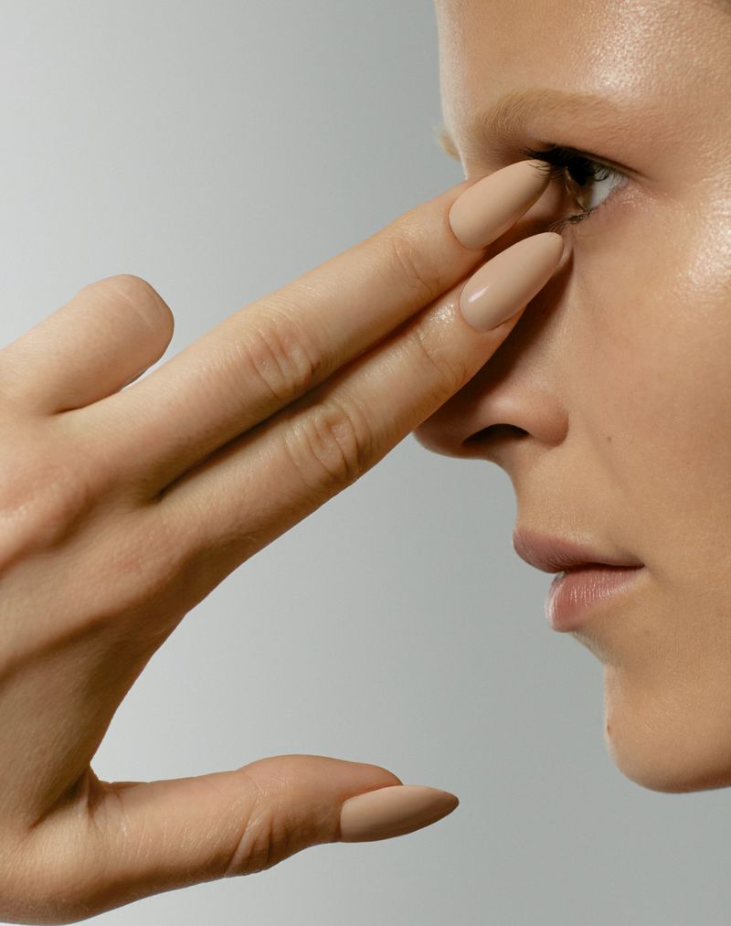Close-up beauty shot of an adult female model with nude makeup and light skin, hand near face, against a light gray background.