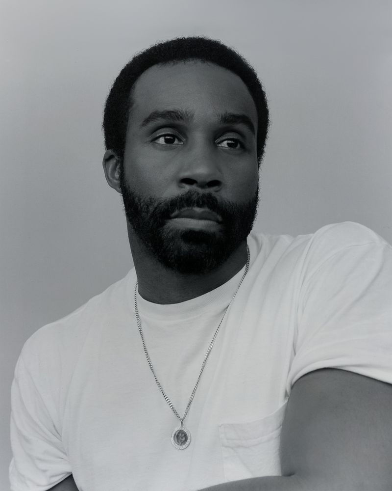 Monochrome portrait of a bearded adult male in a white t-shirt looking away from the camera against a solid grey background.