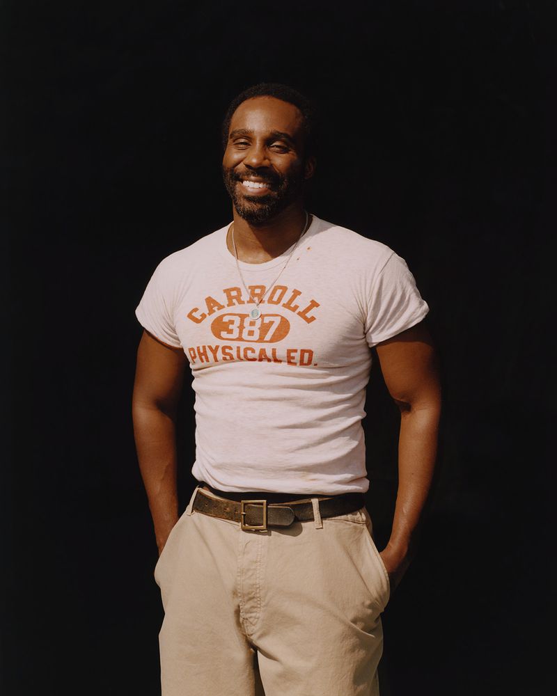 Smiling African-American man in a vintage-style t-shirt and khaki pants stands against a dark backdrop, looking directly at the camera.