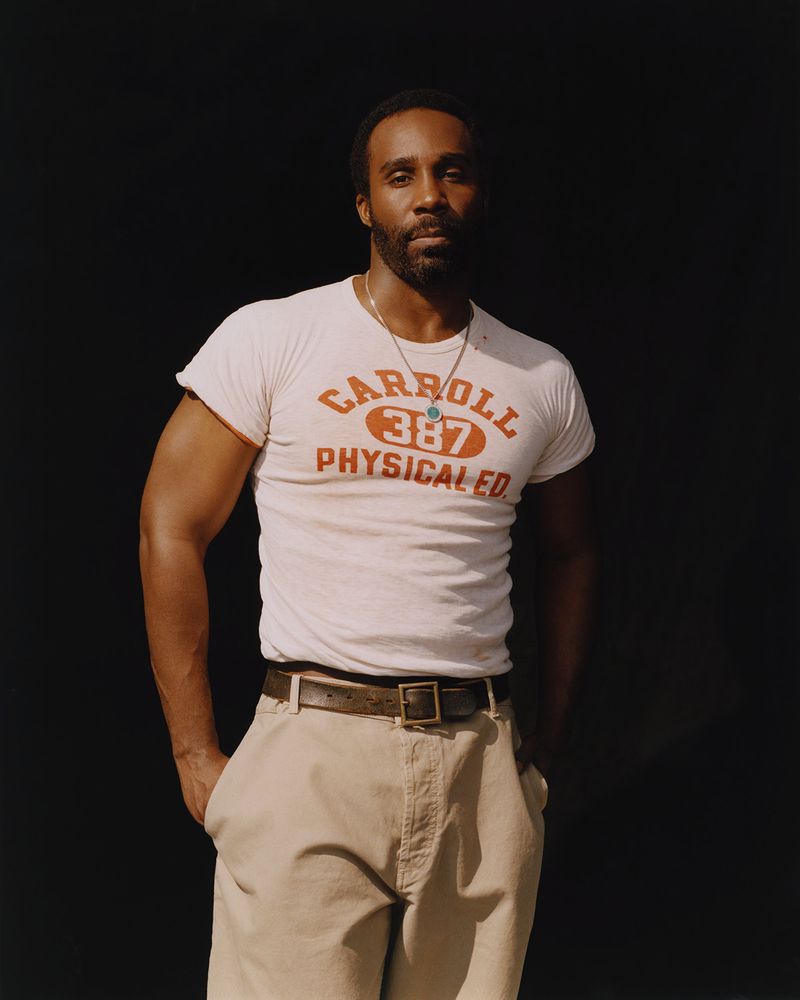 Portrait of an African American man in a t-shirt, standing in front of a dark backdrop in a medium shot, with a neutral facial expression.