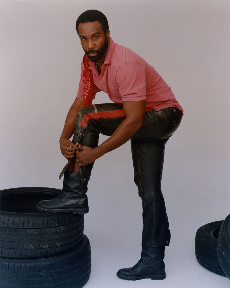 Adult male poses in pink polo, black leather pants and boots, standing on tires in a studio environment with neutral background.