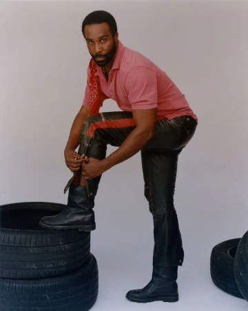Adult male poses in pink polo, black leather pants and boots, standing on tires in a studio environment with neutral background.