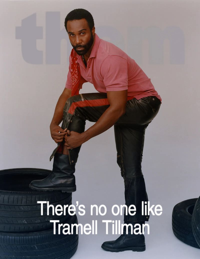 African-American man in black leather pants poses indoors, left leg resting on tires, wearing boots and red polo shirt against white backdrop.