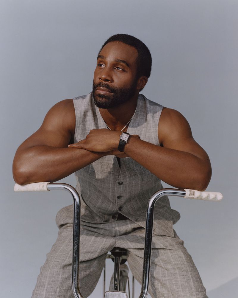 Portrait of a black adult male in vest sitting on a vintage bicycle in a light blue studio setting, gazing to the side, medium shot.