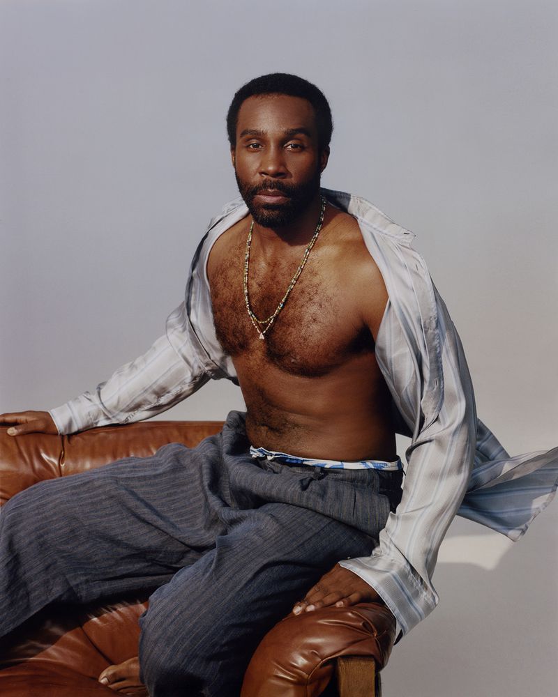 Portrait of a Black man in an unbuttoned shirt and slacks sitting on a brown leather chair against a neutral backdrop.