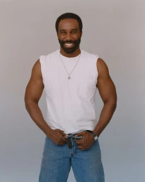 Smiling black man wearing a white t-shirt and blue jeans against a grey studio backdrop for them.us June 2025 Cover.