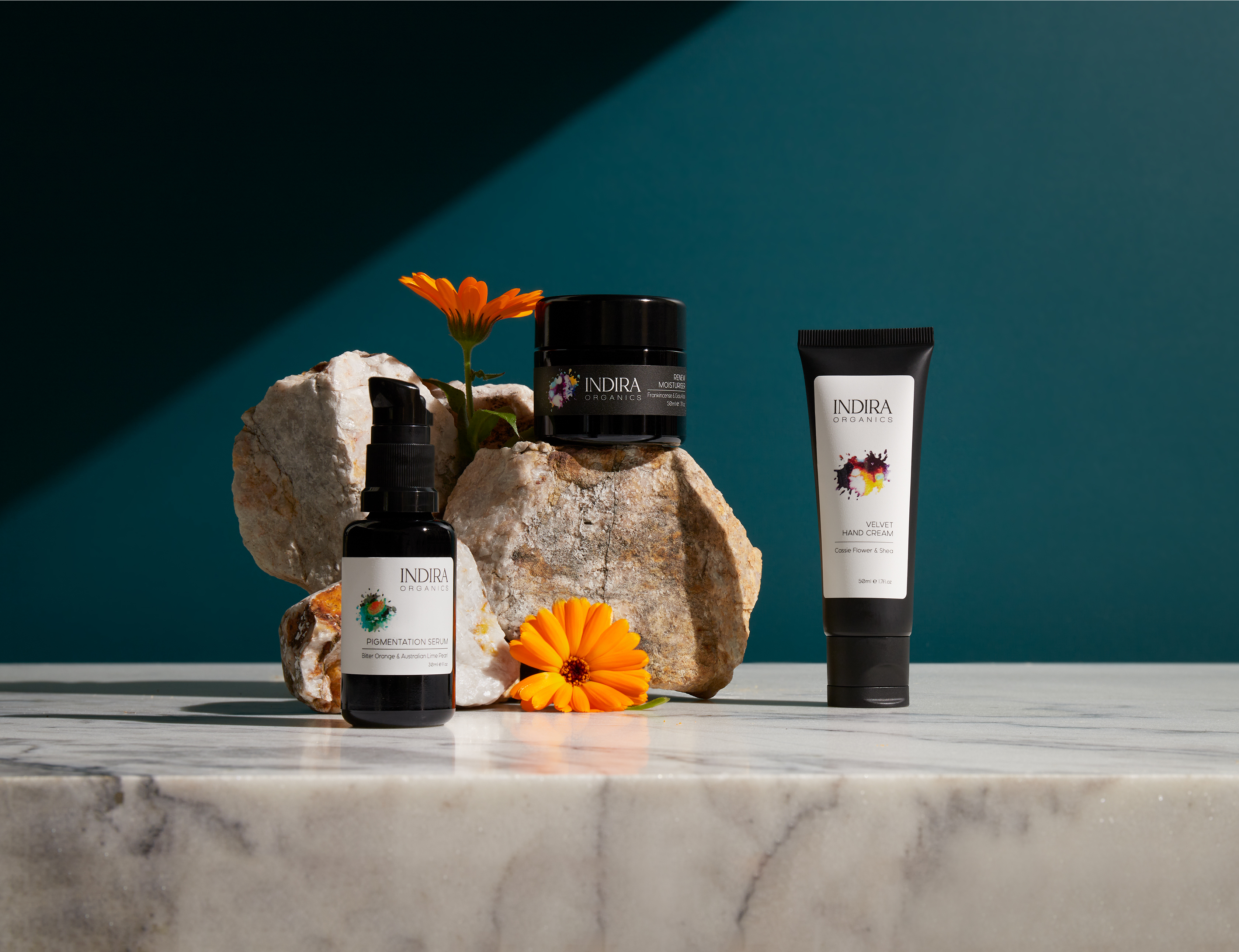 Indira Organics skincare product photography featuring a bottle, jar, and tube on a marble surface with orange flowers and teal background.