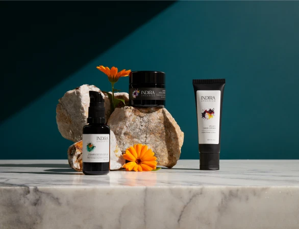 Indira Organics skincare products displayed on white marble rocks with orange flower blossoms against a deep teal background