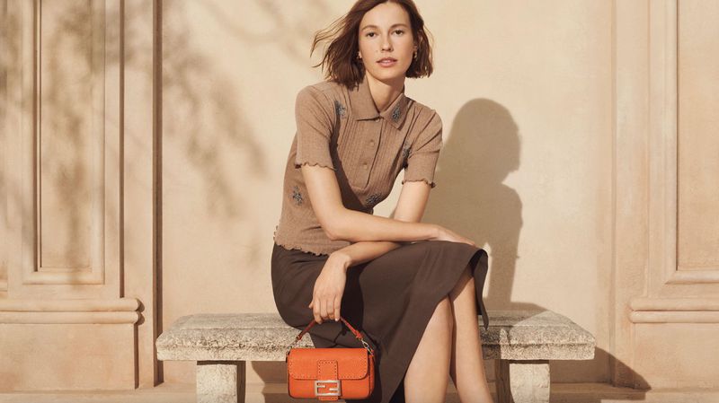 A woman sits on a stone bench, holding an orange handbag with a metal logo, wearing a brown top and skirt, bathed in soft, natural light.