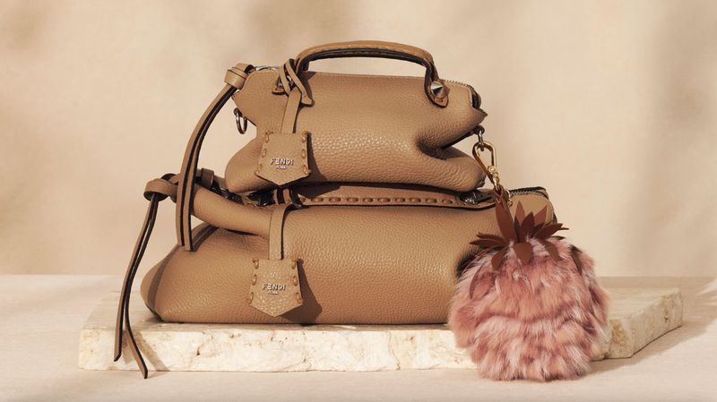 Two light brown Fendi handbags are stacked on a light marble surface with a fur charm on the lower bag. Beige background.