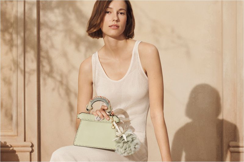 Adult woman poses with a light green satchel handbag and fuzzy charm, wearing a knit top in a studio setting with soft lighting.
