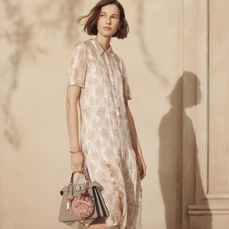 An adult female model wears a button-down dress while holding a beige handbag in a studio environment with a neutral backdrop.