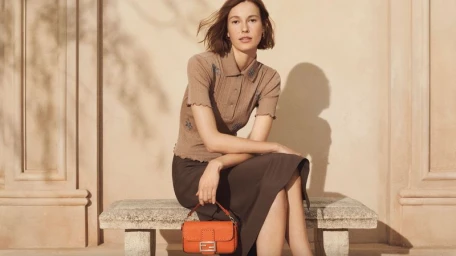 Model seated on a stone bench wearing a brown polo shirt and holding an orange leather handbag against a wall.
