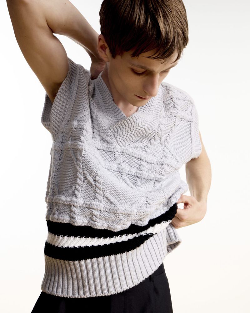 A person wearing a knitted sweater vest in front of a white background, with their right arm raised behind their head.