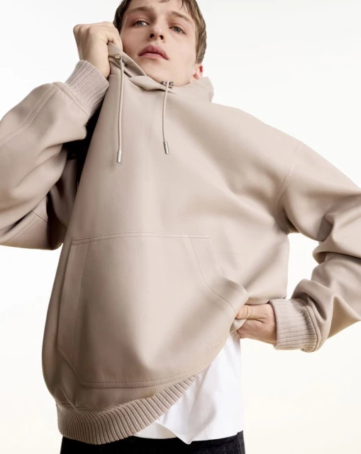 A male model poses indoors wearing a tan hooded sweatshirt with a pocket. He is partially cropped and looks at the camera.