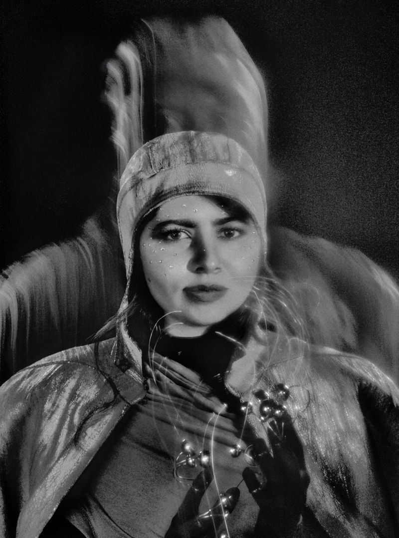 Black and white portrait of a woman with a hat holding a lit string of lights, featuring a blurred figure in the background.