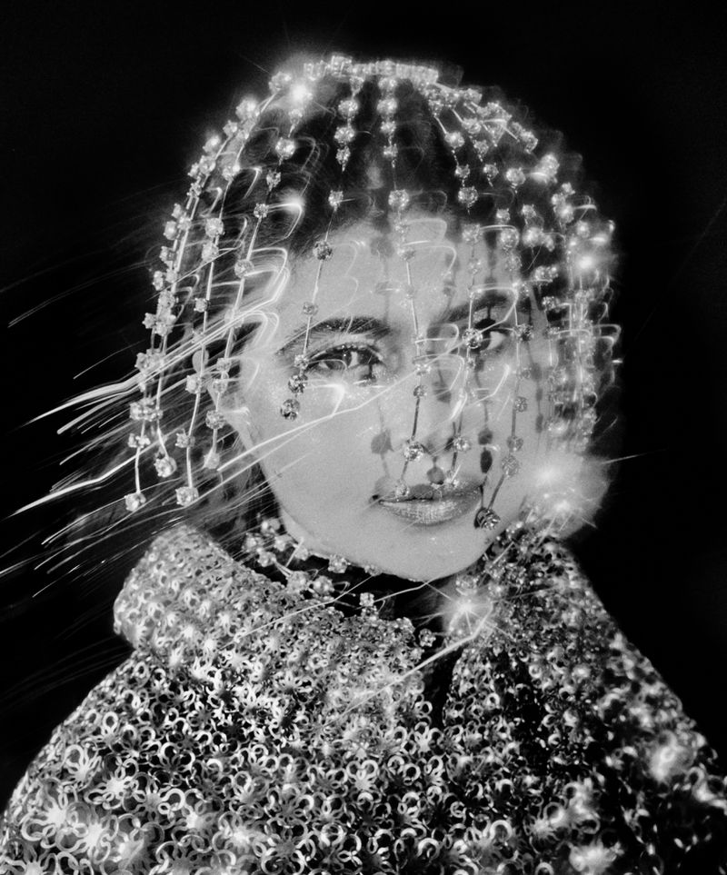 Monochrome portrait of a woman wearing an ornate beaded headpiece and decorative collar, set against a dark background. High-fashion glamour shot.