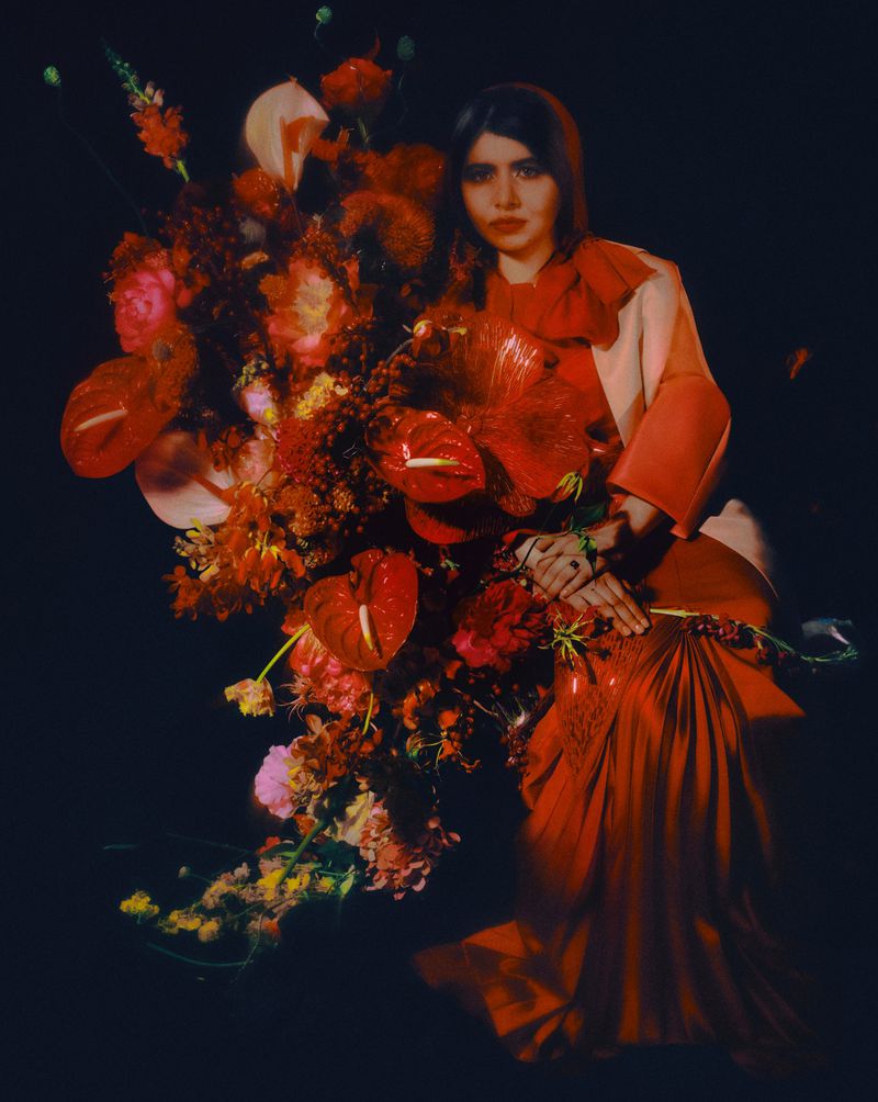 Woman in red dress and flowers against a dark background. Portrait shot featuring floral arrangement and fashion elements. Editorial style.