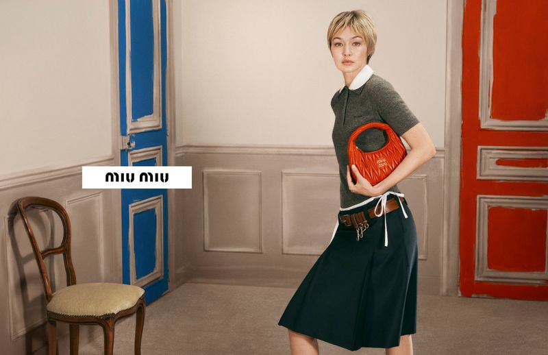 Gigi Hadid posing in a grey polo shirt and dark skirt holding an orange bag in an interior with blue and red doors.
