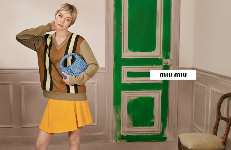 Fashion shot of a woman holding a blue Miu Miu purse, wearing a brown sweater, and yellow skirt next to a painted green door.