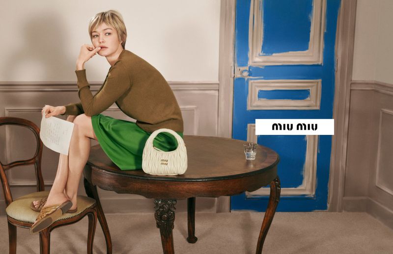 Model Gigi Hadid sitting on a round wooden table holding a letter, featuring a white Miu Miu leather handbag
