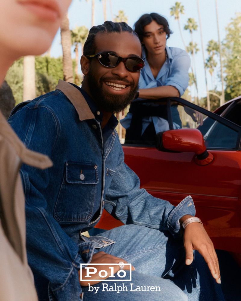 Two men pose next to a red convertible on a palm tree-lined street. One man in denim smiles at the camera. Fashion and automotive.