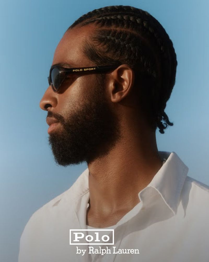 Profile portrait of a Black man with cornrows wearing sunglasses and a white collared shirt against a clear blue sky