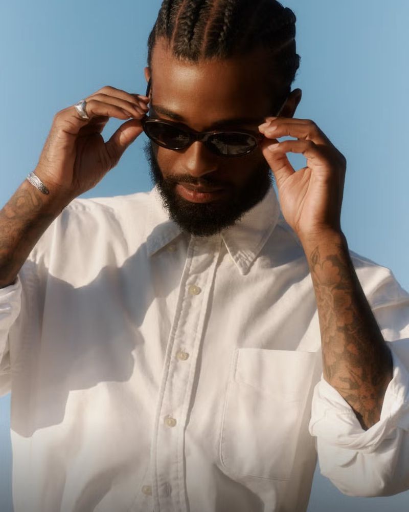 Man wearing a white shirt and sunglasses adjusting his eyewear against a clear blue sky background
