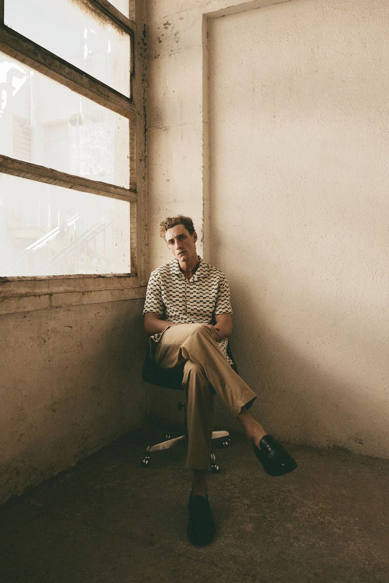 Model seated on a chair next to a large window wearing a patterned short-sleeve shirt and khaki trousers.