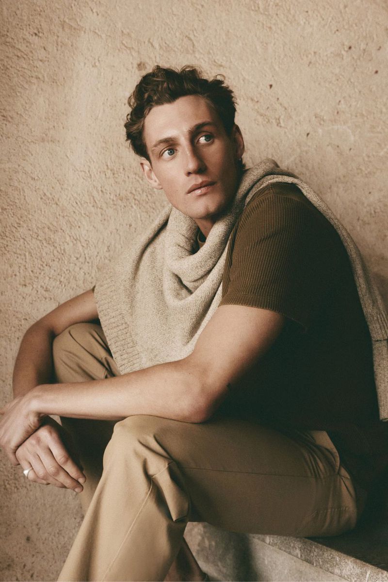 Male model wearing a brown t-shirt and tan trousers with a cream sweater draped over his shoulders against a textured wall.