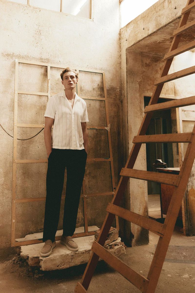 Male model in a white short-sleeve shirt and dark trousers standing inside an unfinished rustic building with a wooden ladder.