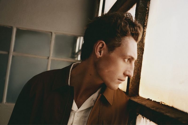 Profile view of a man with curly hair wearing a brown jacket looking out a large gridded window for Sfera S/S 26