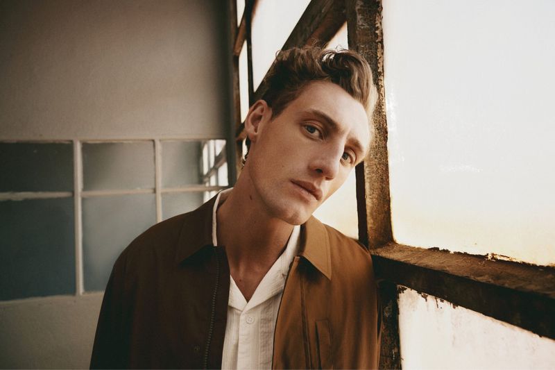 Close-up portrait of a male model leaning against a window frame wearing a brown jacket over a white shirt.