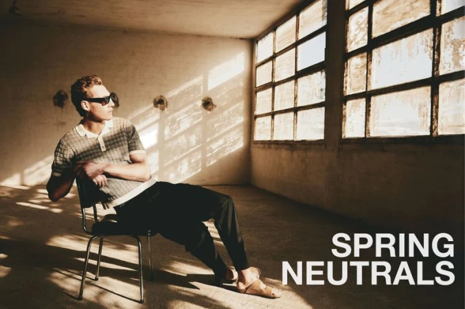 A man wearing sunglasses, a knit polo shirt, and trousers seated on a chair inside a rustic room with grid windows casting dramatic shadows.