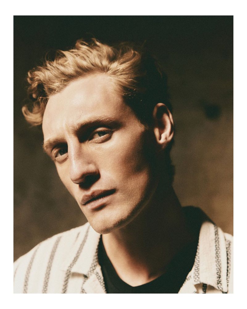 Close-up portrait of a male model with blond hair wearing a striped shirt and dark t-shirt against a dark background