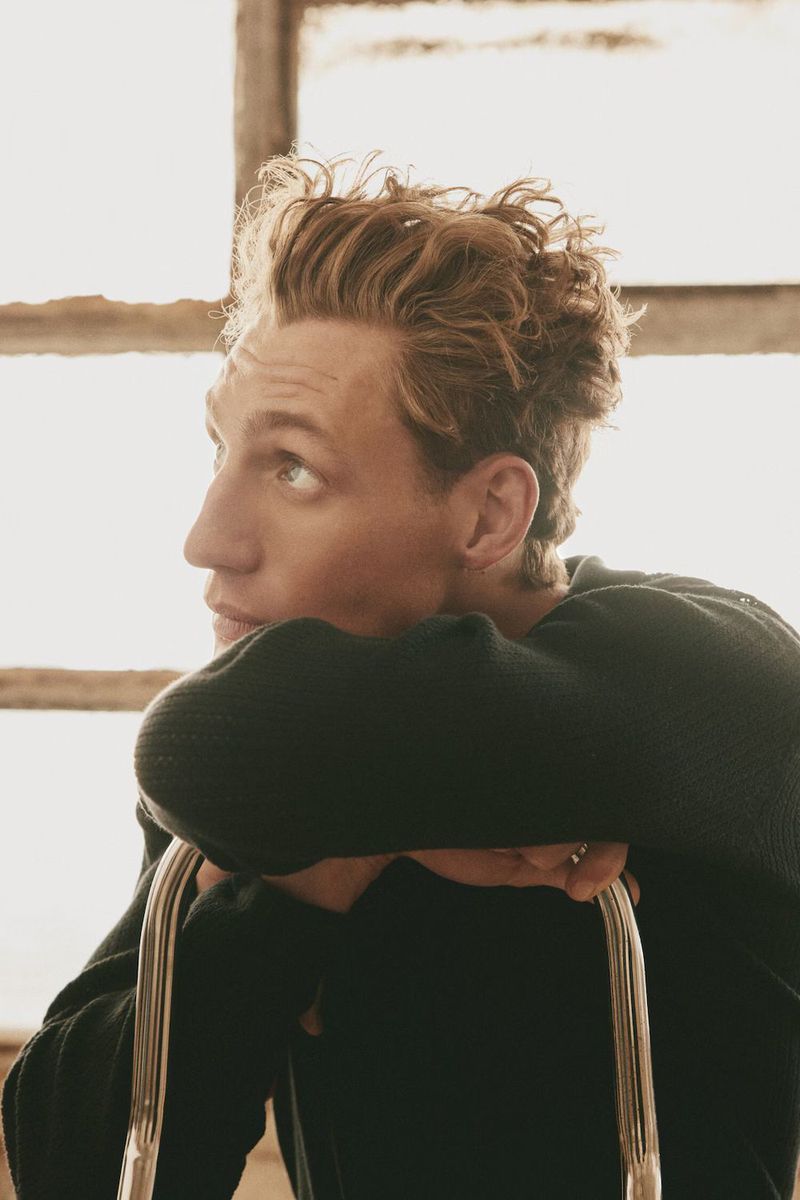 Portrait of a young man with curly blonde hair wearing a black sweater, resting his arms on a chair back.