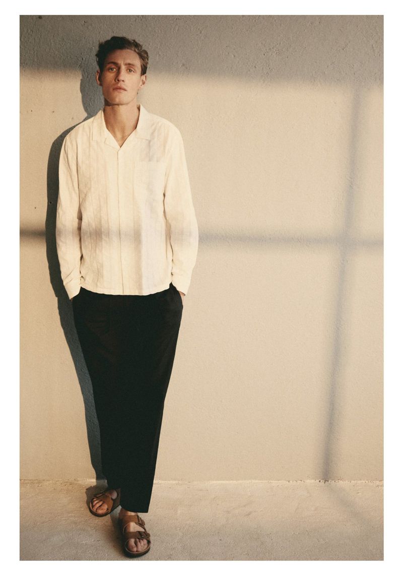 A full shot of a male fashion model in an off-white long-sleeved shirt, black pants, and sandals against a beige wall with a shadow.
