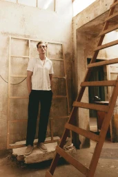 Male model in a white short-sleeve shirt and dark trousers standing inside an unfinished rustic building with a wooden ladder.