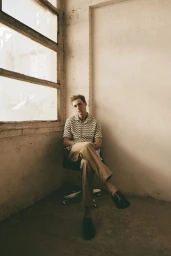 Model seated on a chair next to a large window wearing a patterned short-sleeve shirt and khaki trousers.